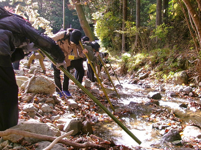 金目川源流の浅い川の流れに、竹の筒をさし、筒に耳を当てて源流のせせさぎを聴く参加者たち